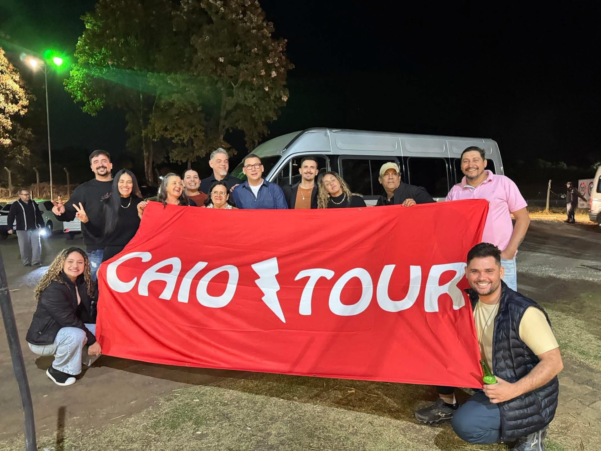 Grupo do Caio Tour à noite ao lado da van, segurando a faixa